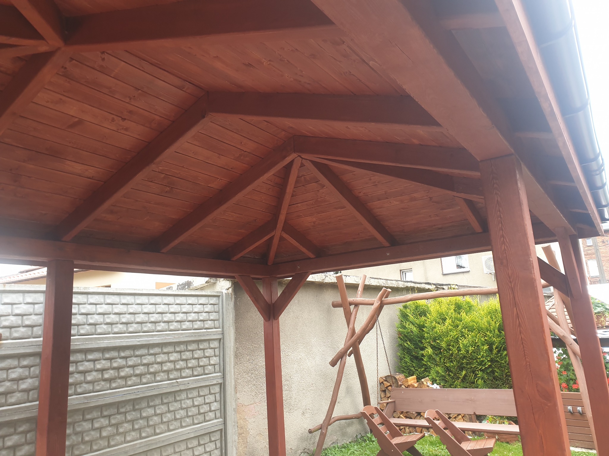 Drewniana altana ogrodowa z widoczną konstrukcją dachu, pomalowana na brązowo, z fragmentem rynny i betonowym ogrodzeniem w tle.