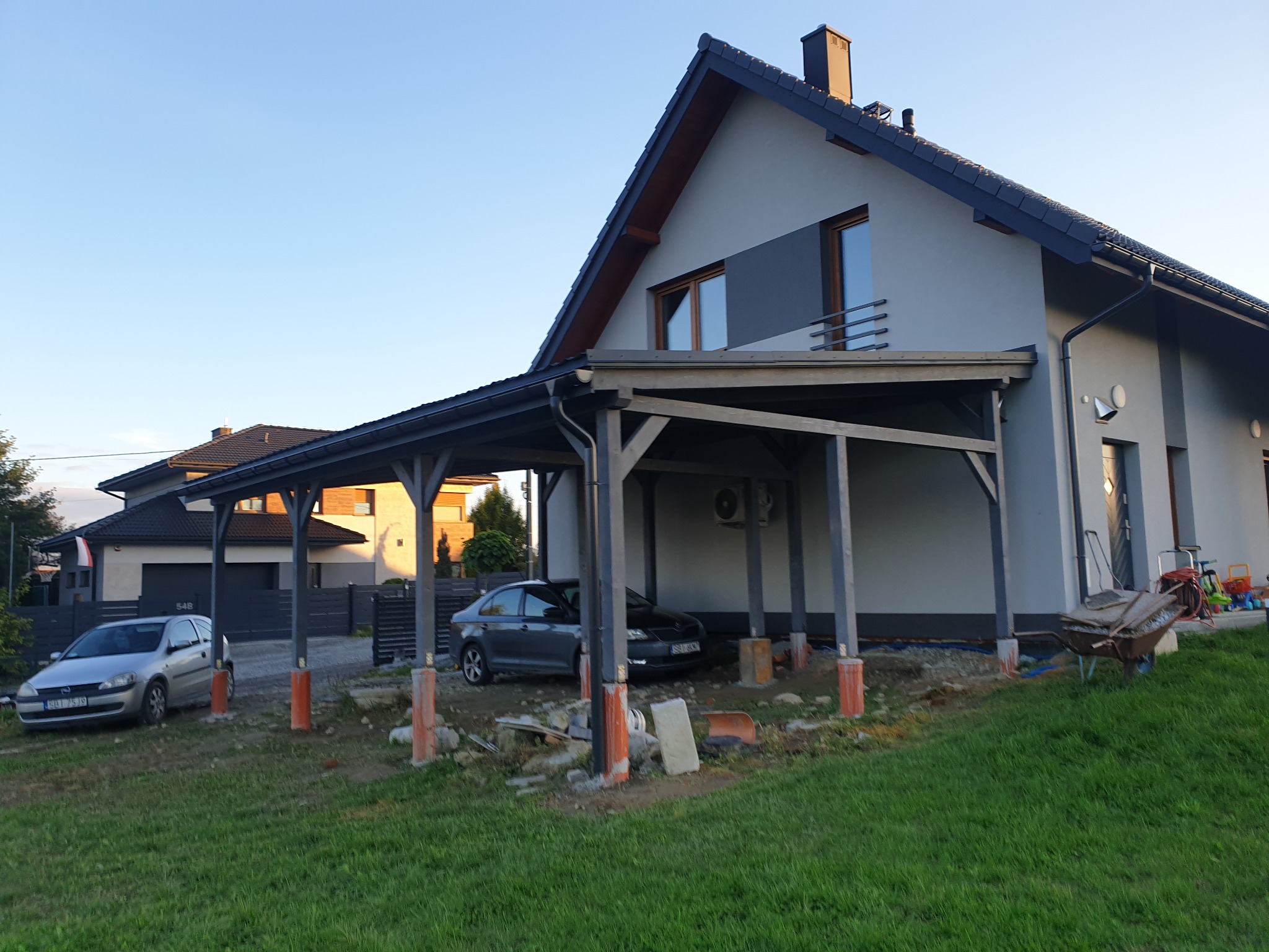 Nowoczesny dom z szarą elewacją i drewnianą konstrukcją zadaszenia na dwa samochody, widoczne dwa auta, teren wokół w trakcie prac wykończeniowych.