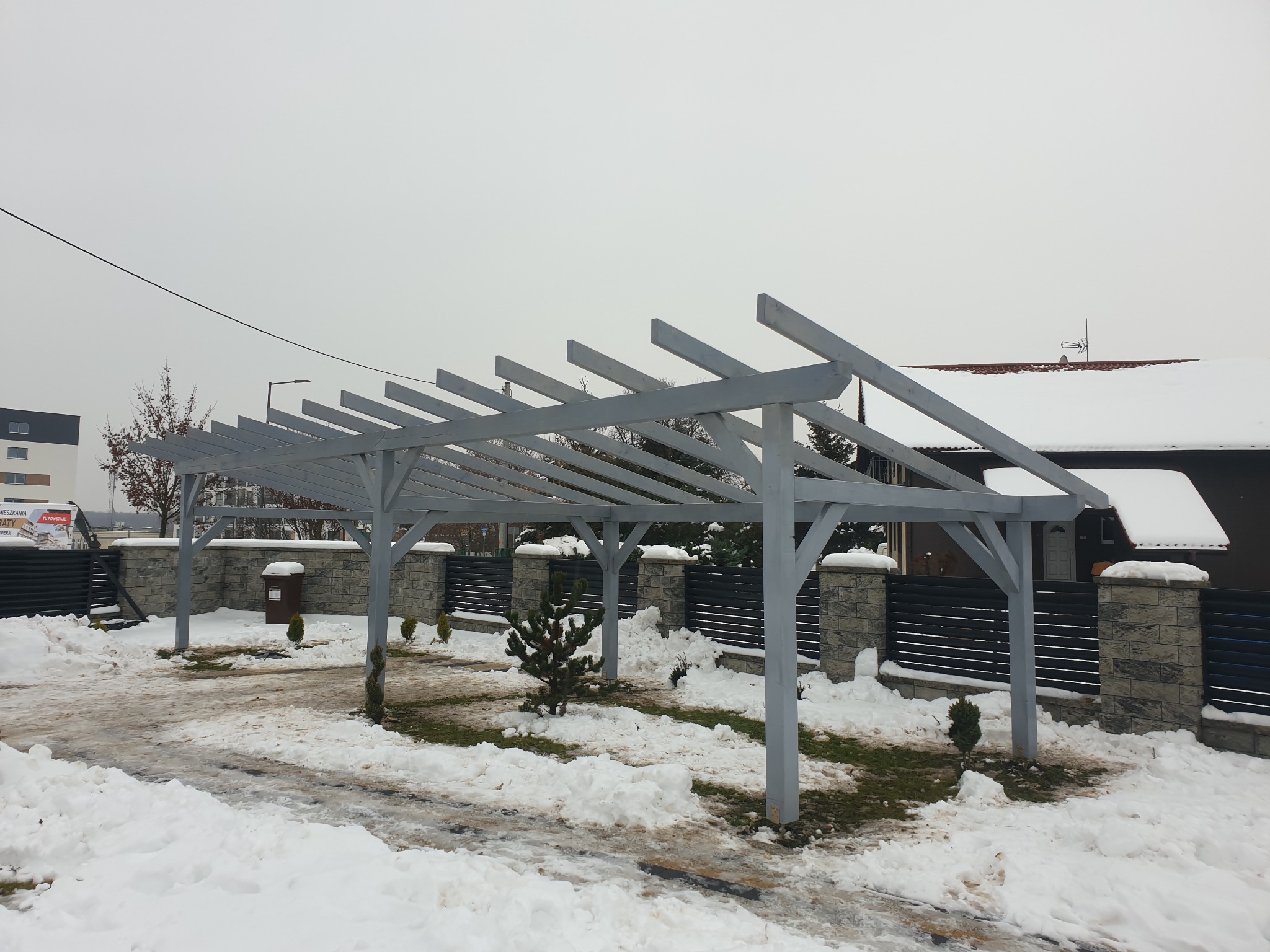 Szara, drewniana konstrukcja zadaszenia tarasu lub wiaty samochodowej w zimowej scenerii, częściowo pokryta śniegiem, z widocznym ogrodzeniem z kamiennymi słupkami i drewnianymi panelami w tle.