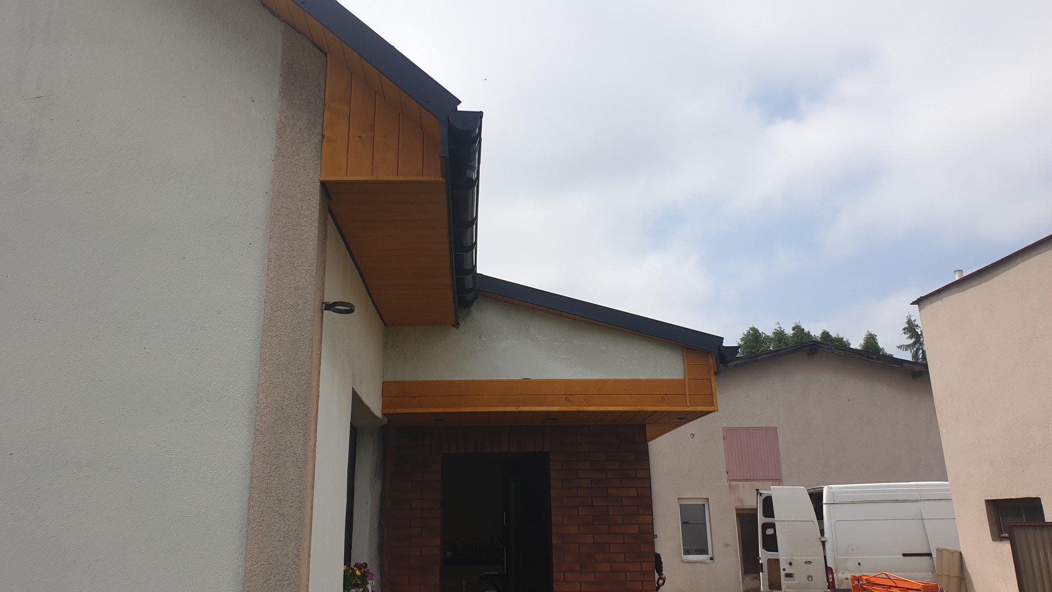 Fragment domu z drewnianą podbitką dachu, czarnymi rynnami i ceglanym wejściem, widok z poziomu gruntu na tle pochmurnego nieba i zaparkowanego białego vana.