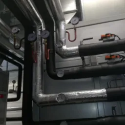 Rzędy rur instalacyjnych, niektóre izolowane folią aluminiową, z zamontowanymi manometrami, w pomieszczeniu technicznym.