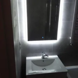 Umywalka z szafką i podświetlanym lustrem w nowoczesnej łazience wykończonej szarymi płytkami, w tle widoczna kabina prysznicowa.