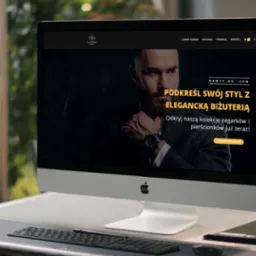 Ekran komputera iMac prezentuje stronę internetową sklepu z biżuterią, na pierwszym planie klawiatura i tablet graficzny na metalowym stole.