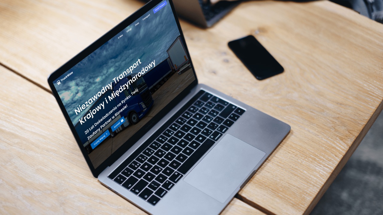 Laptop wyświetla stronę internetową firmy transportowej TruckWaw z hasłem 'Niezawodny Transport Krajowy i Międzynarodowy', smartfon obok na drewnianym blacie biurka.