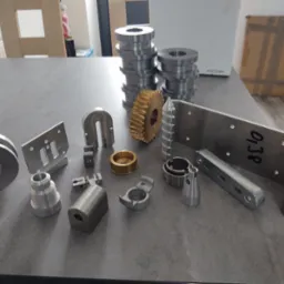 Różnorodne metalowe detale i komponenty, w tym przekładnia zębata, tuleje, elementy z otworami i gwintami, ułożone na szarym blacie w neutralnym oświetleniu.
