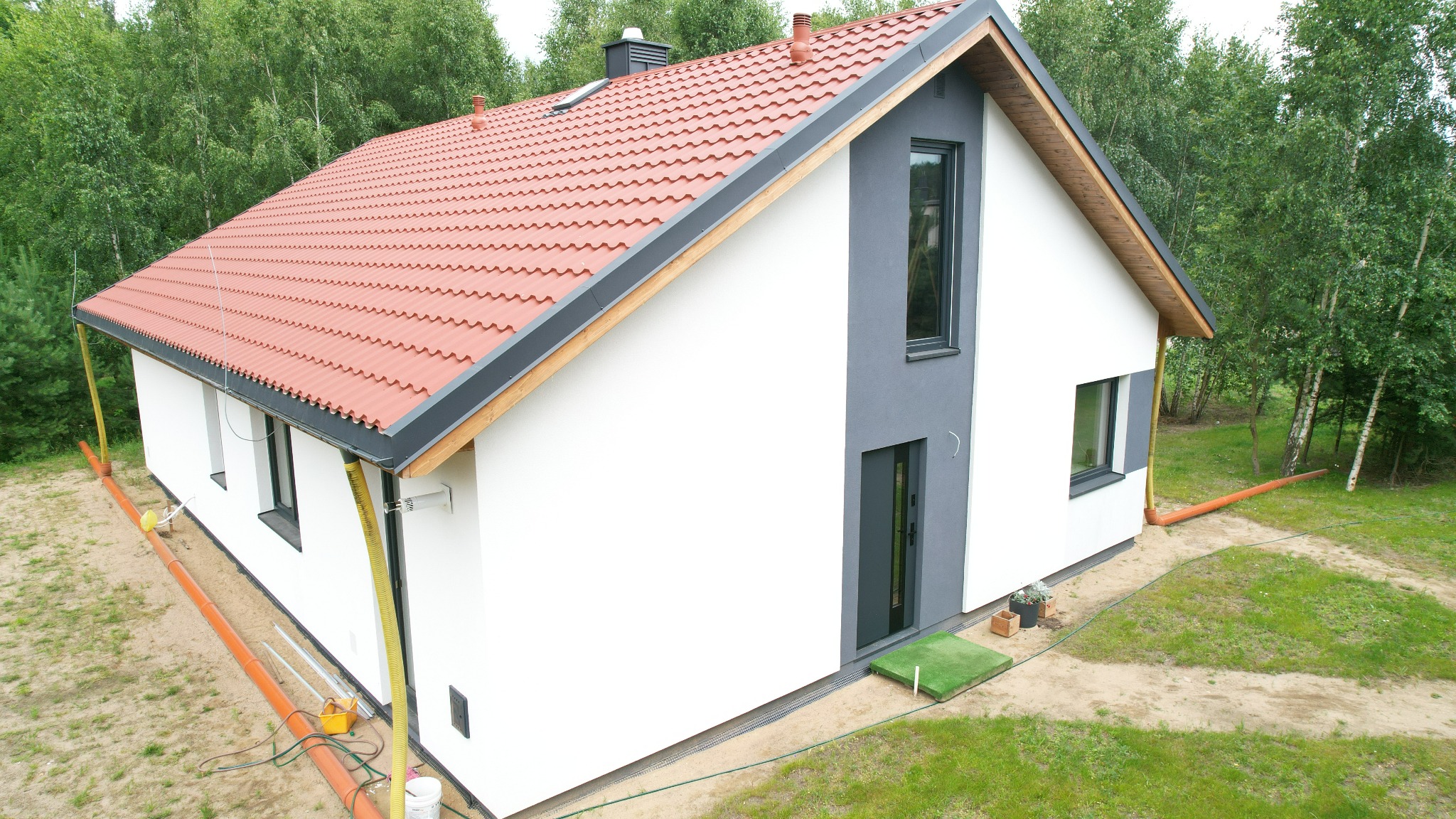 Nowoczesny dom z dachem z czerwonej dachówki, białą elewacją z szarymi akcentami i widocznymi rurami drenażowymi, otoczony zielenią.