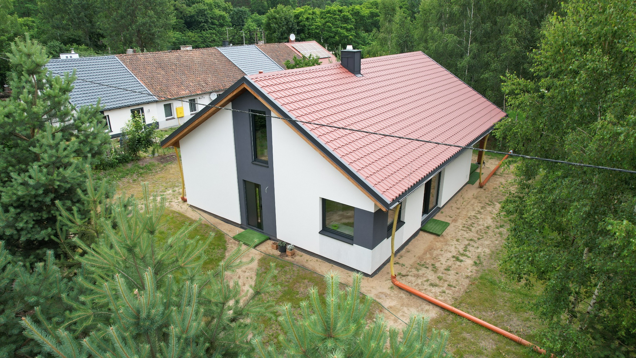 Nowoczesny parterowy dom z czerwoną dachówką, białą elewacją i szarymi akcentami, otoczony zielenią. Widok z góry, ukazujący ukończony budynek z elementami wykończenia.