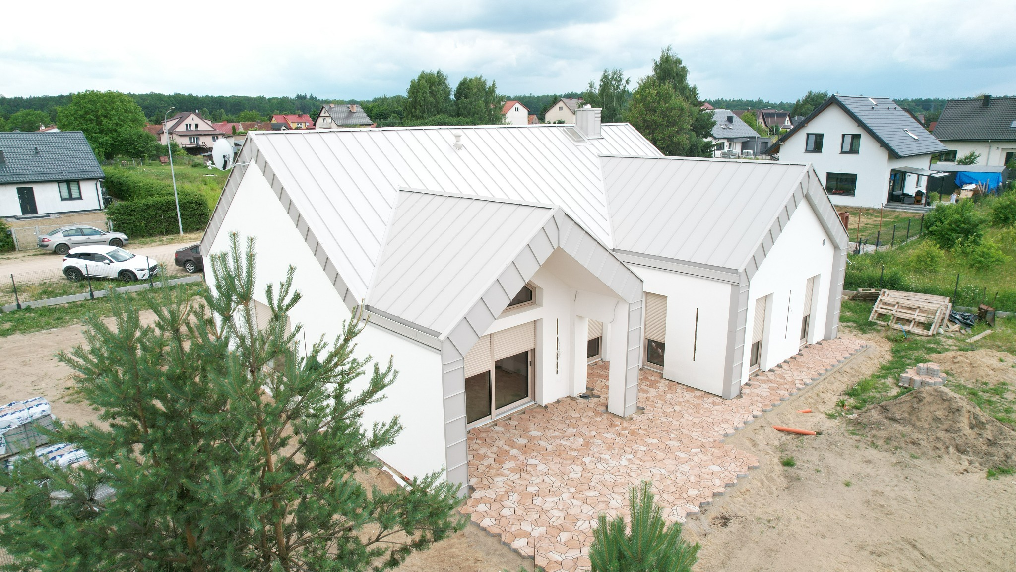 Nowoczesny dom z białymi ścianami i szarym dachem z blachy na tle zieleni. Uwagę przykuwa wzór kostki brukowej na podjeździe i elementy wykończeniowe.