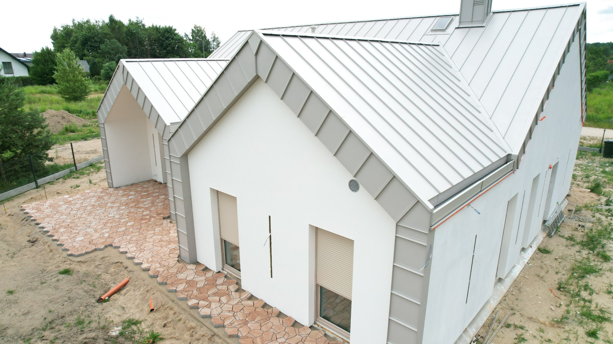 Nowoczesny dom z białymi ścianami i szarym dachem z blachy na rąbek stojący, z częściowo ułożoną kostką brukową na podjeździe i odsłoniętym gruntem.
