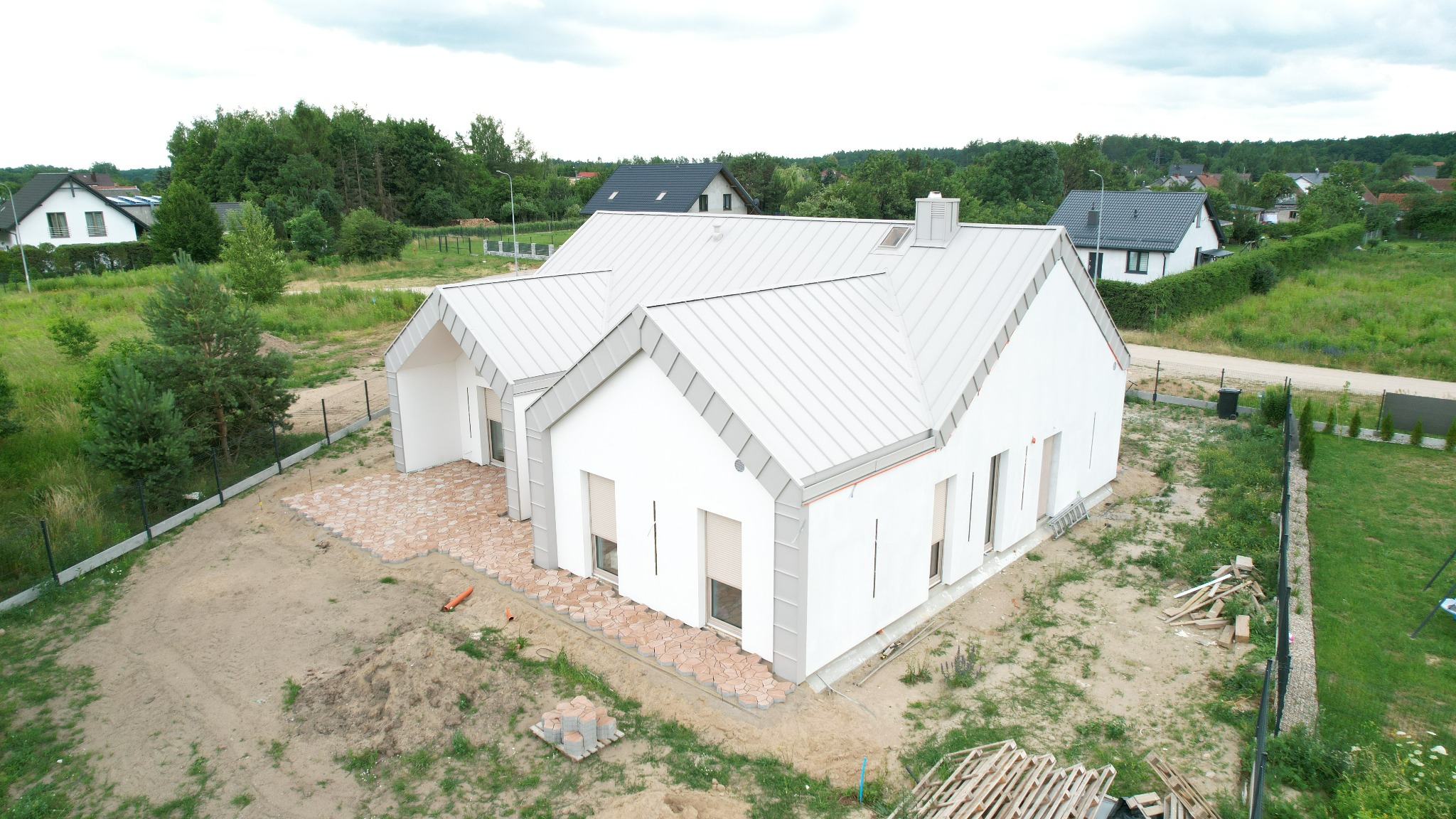 Nowoczesny, biały dom z szarym dachem z blachy na etapie wykończenia. Widok z góry na budynek i teren wokół z kostką brukową. W tle zieleń i inne domy.