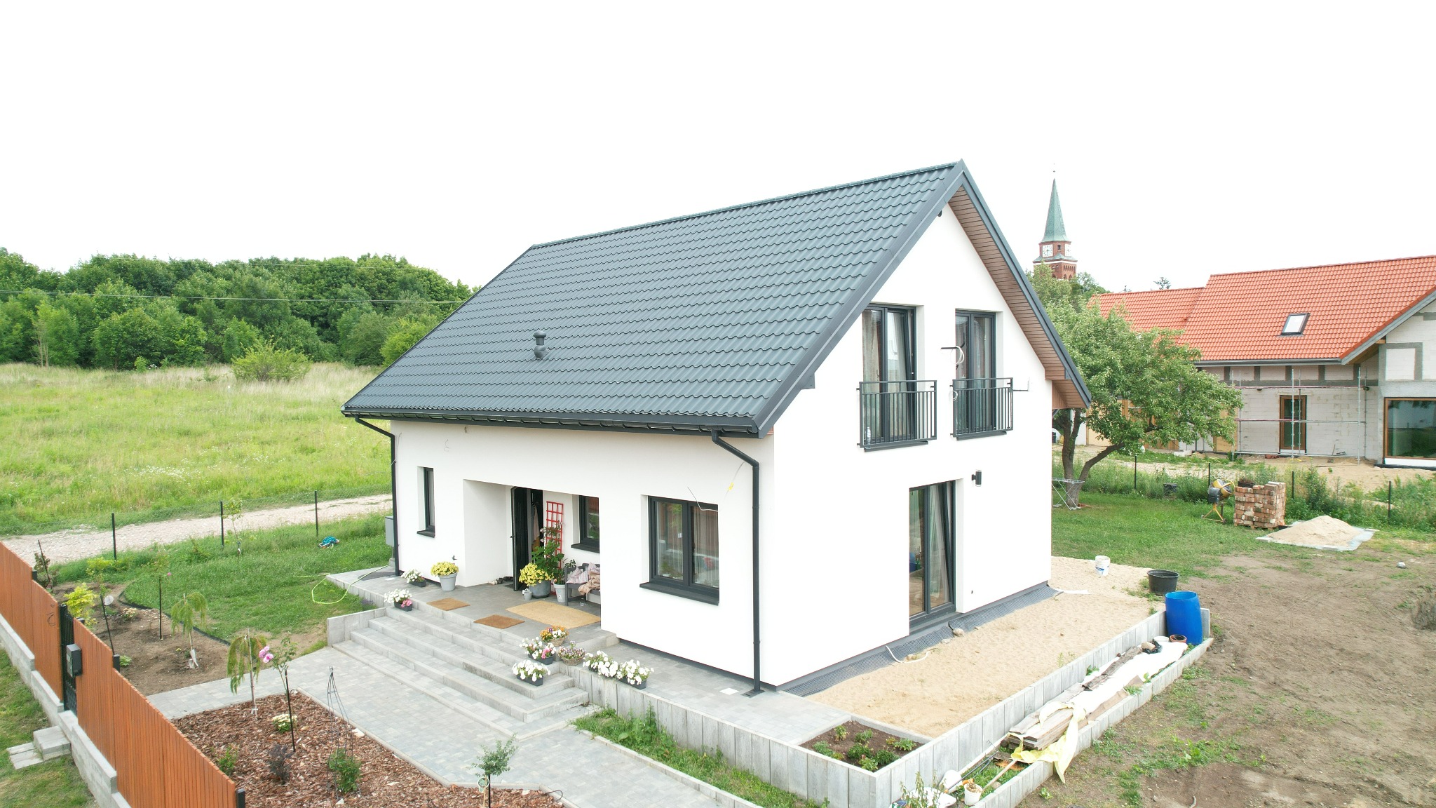 Nowoczesny, biały dom z szarym dachem dwuspadowym, balkonami i brukowanym podjazdem. W tle widoczny kościół i inne budynki.