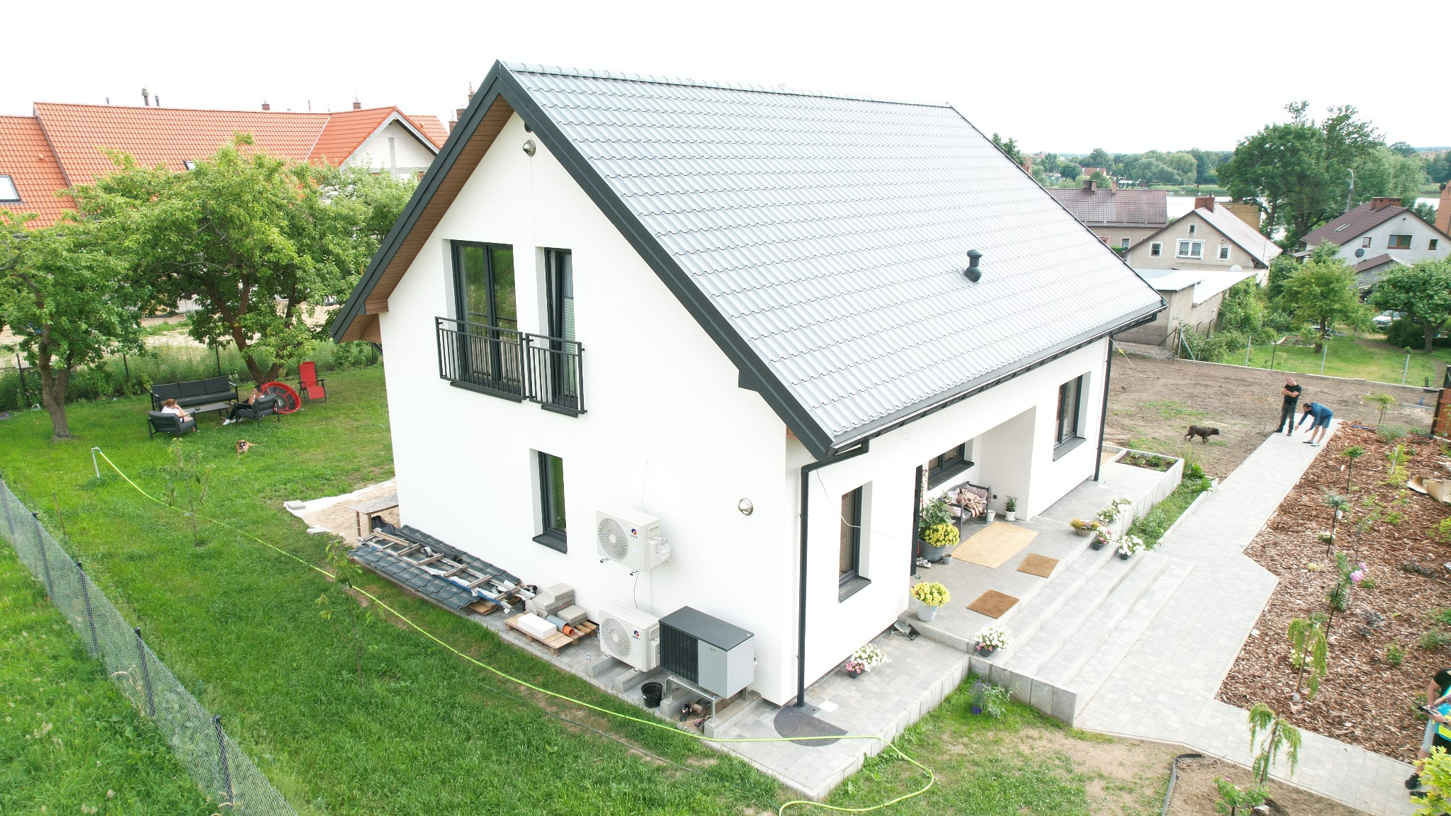 Nowoczesny, biały dom z szarym dachem z blachodachówki, balkonem i klimatyzacją. Widok z góry na zagospodarowany teren wokół budynku.