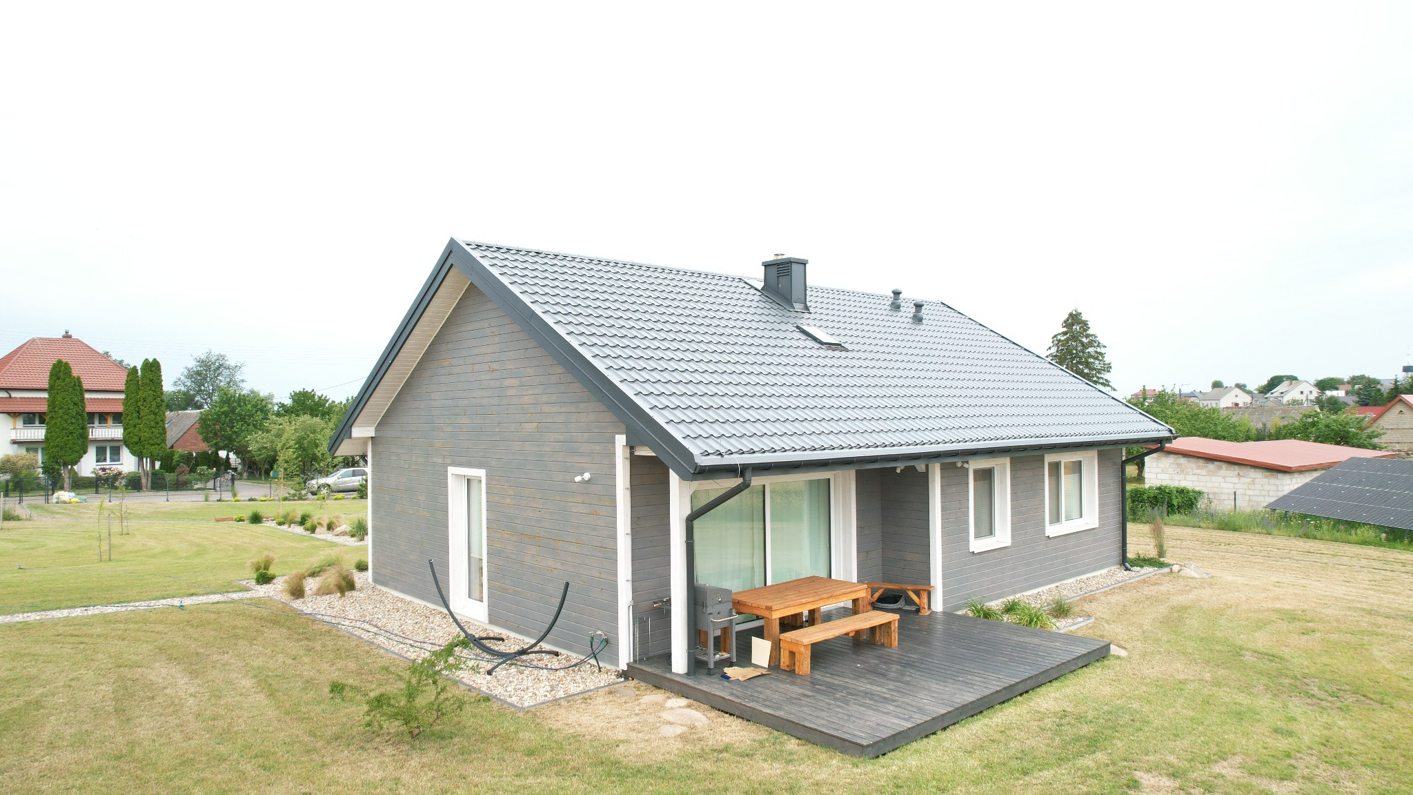 Nowoczesny dom szkieletowy z szarą elewacją i drewnianym tarasem z meblami ogrodowymi. Widok z góry na budynek i otaczający go trawnik.