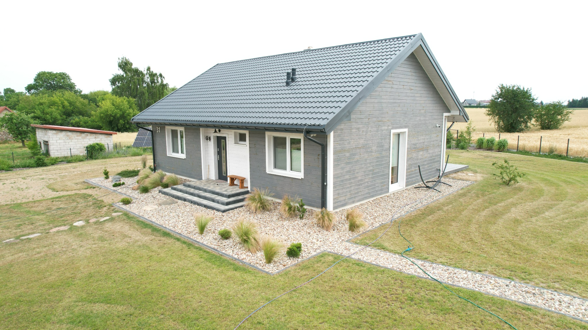 Parterowy dom z szarą elewacją drewnianą i ciemną dachówką, otoczony trawnikiem i ozdobnymi kamieniami. Widoczne panele solarne w tle i mała ławeczka przy wejściu.