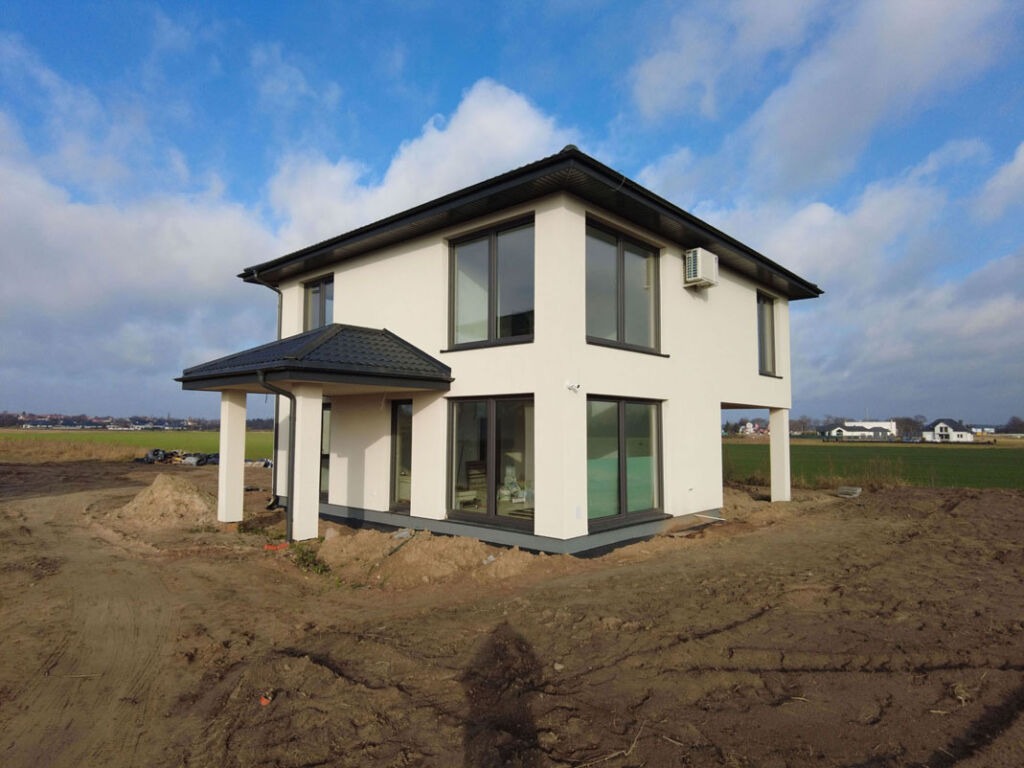 Dwukondygnacyjny dom z płaskim dachem i przeszklonymi drzwiami balkonowymi, zadaszone wejście wsparte na kolumnach, klimatyzator na elewacji, otoczony polem i widocznymi w oddali zabudowaniami.