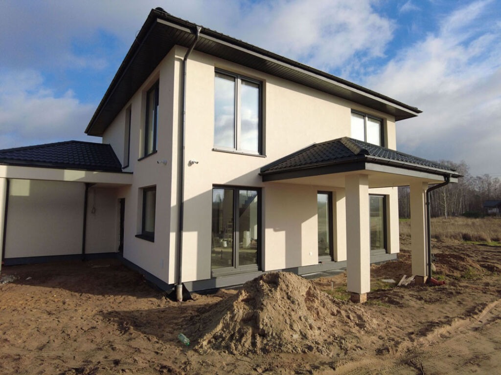 Dwukondygnacyjny dom z jasną elewacją i ciemnym dachem, widoczny z zewnątrz, z przygotowanym terenem wokół budynku.