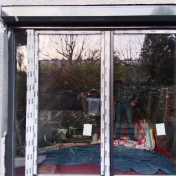 Wojt-Plast - Montaż dwuskrzydłowego okna PCV z widocznym refleksyjnym odbiciem dwóch osób w trakcie pracy i narzędziami wewnątrz pomieszczenia, zabezpieczonego folią.