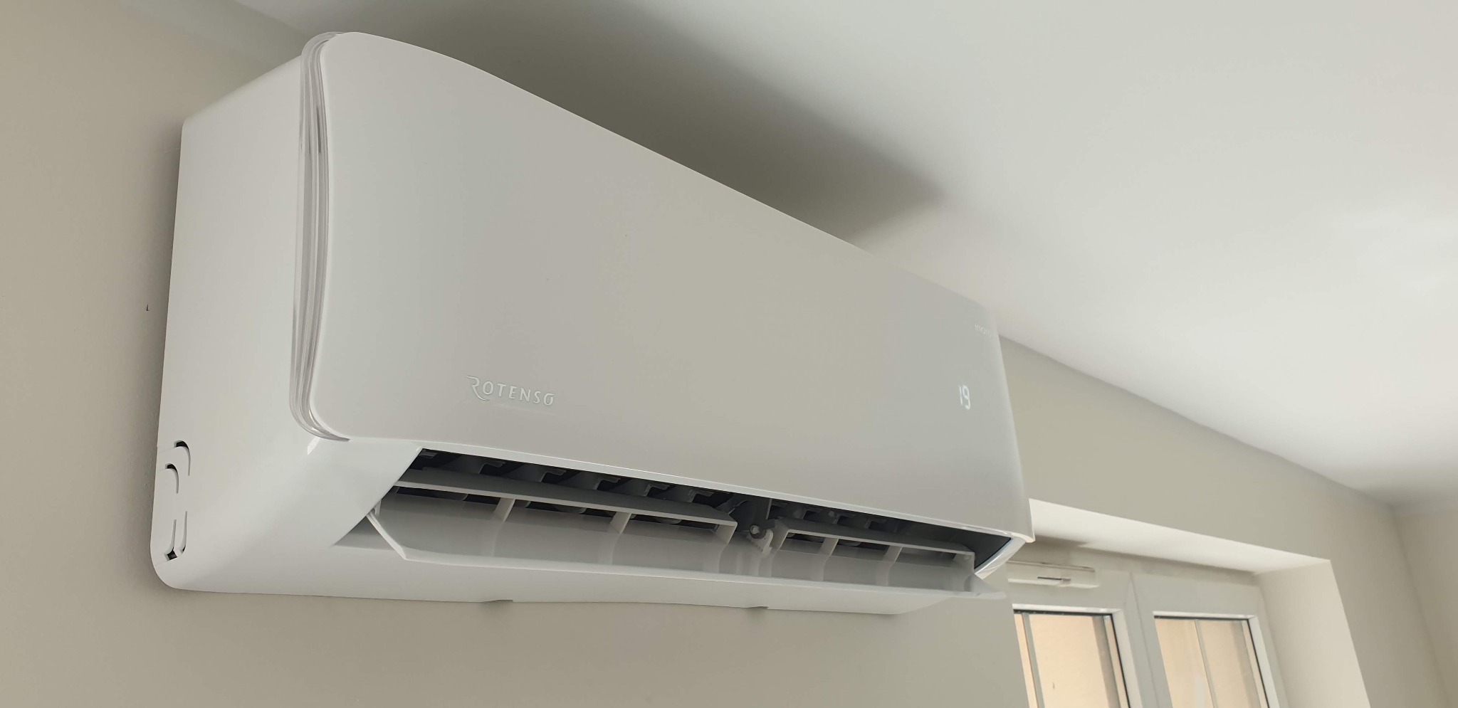 Biały klimatyzator marki Rotenso zamontowany na ścianie w pomieszczeniu z oknem, widoczny wyświetlacz z temperaturą 19 stopni.