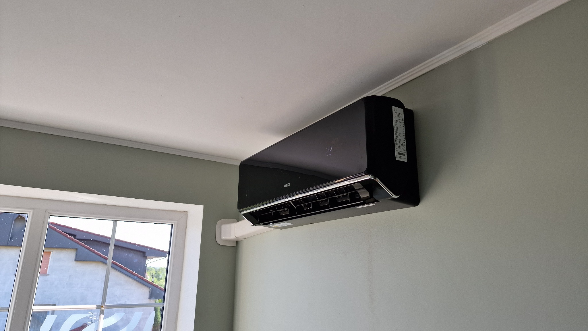 Czarna jednostka klimatyzacji AUX zamontowana na zielonej ścianie w pokoju z widokiem na dom przez okno.