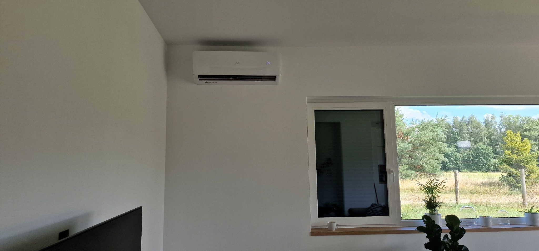 Biały klimatyzator ścienny marki AUX w nowoczesnym salonie z widokiem na zieleń przez okno.