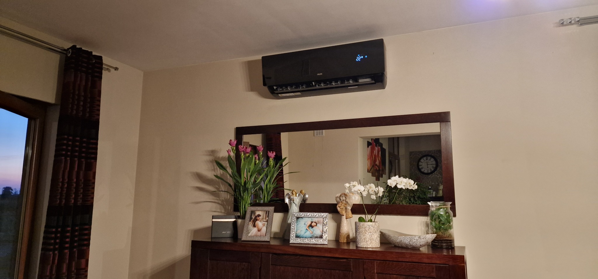 Czarny klimatyzator ścienny z wyświetlaczem temperatury 22 stopnie Celsjusza, zamontowany nad komodą z lustrem i dekoracjami w salonie.