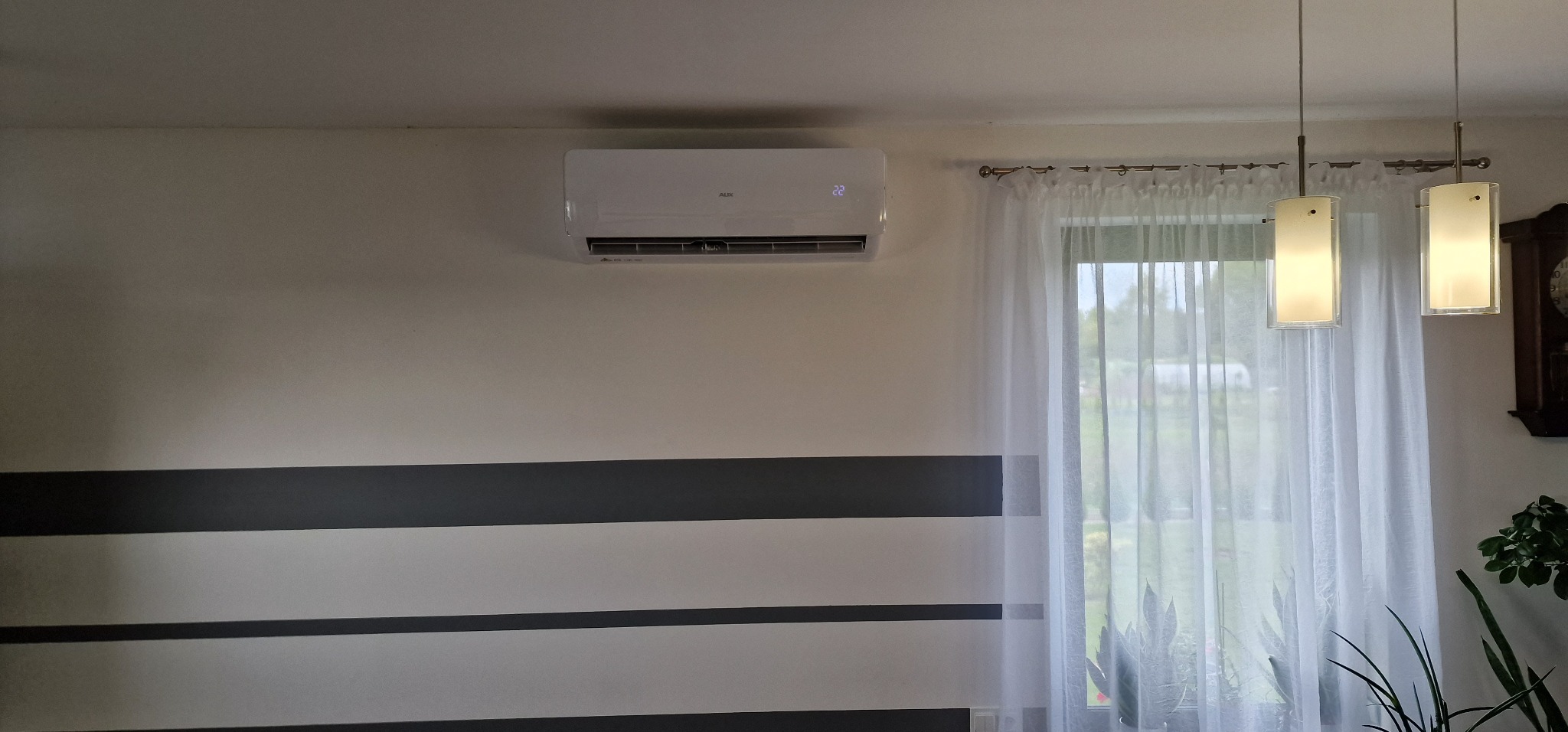 Biały klimatyzator ścienny marki AUX zamontowany w pokoju z poziomymi, szarymi pasami na ścianie, widoczne wiszące lampy i okno z firanką.