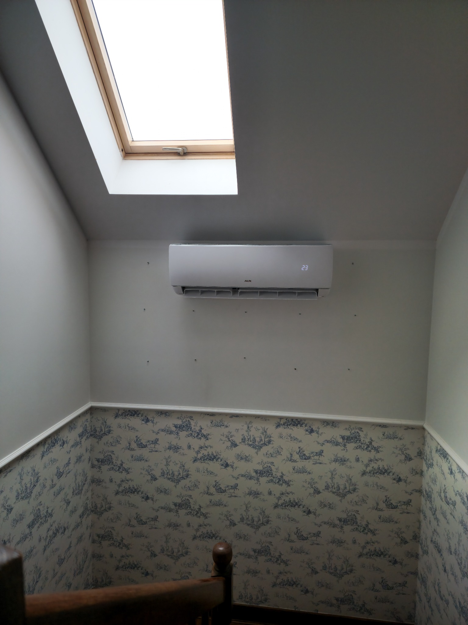 Klimatyzator ścienny marki AUX zamontowany w pokoju na poddaszu, widoczne otwory po poprzednim mocowaniu, ściany wykończone tapetą w stylu rustykalnym, okno dachowe w suficie.