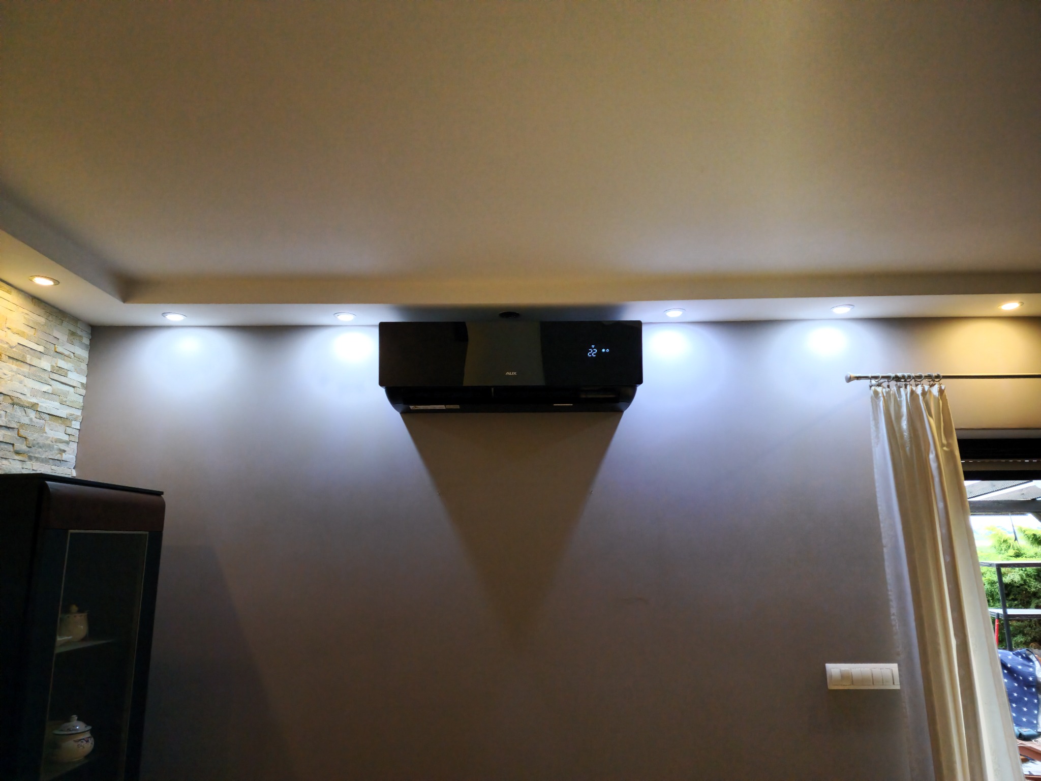 Czarny klimatyzator ścienny marki AUX zamontowany w salonie z szarą ścianą, oświetleniem punktowym i fragmentem kamiennej ściany z lewej strony.