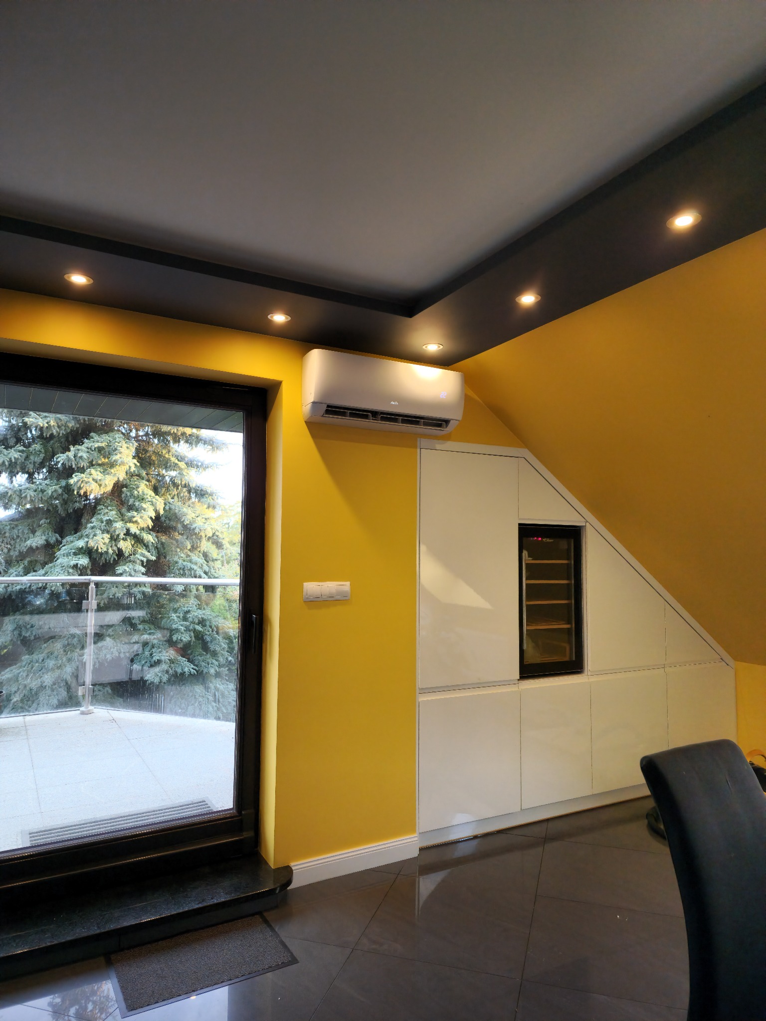 Wnętrze poddasza z klimatyzatorem ściennym, żółtą ścianą, zabudową z szafkami i przeszklonym balkonem z widokiem na drzewo.