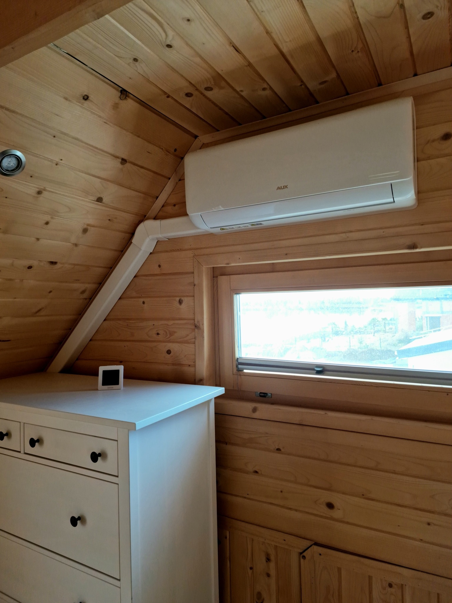 Wnętrze pokoju wykończonego drewnem z zamontowanym klimatyzatorem AUX nad oknem i białą komodą z czarnymi uchwytami.