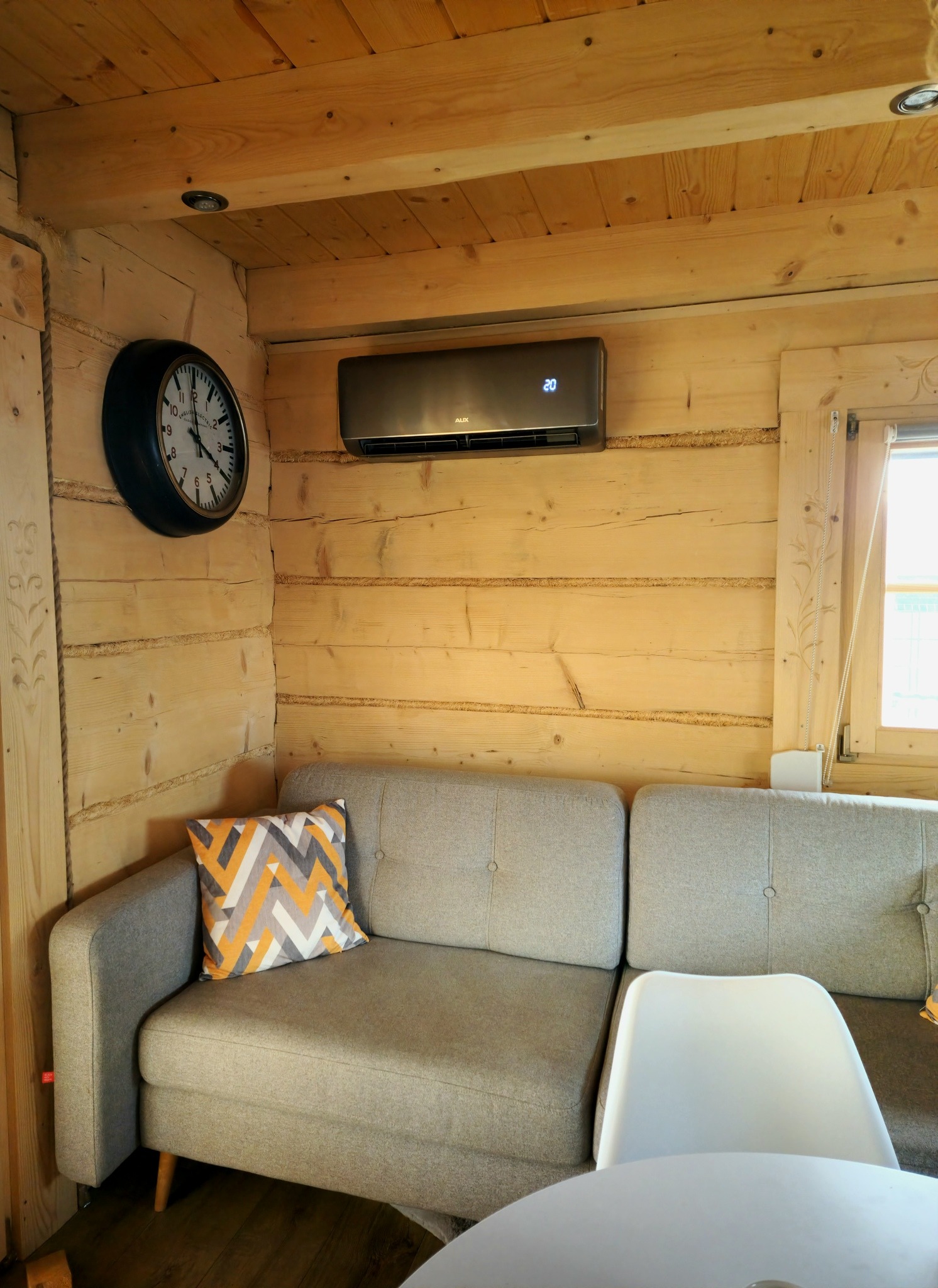 Wnętrze drewnianego domku z szarą sofą, zegarem ściennym i zamontowanym klimatyzatorem z wyświetlaczem temperatury.