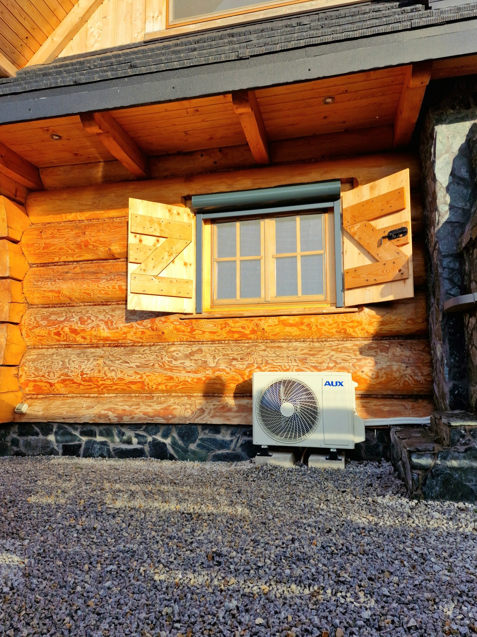 Biała jednostka zewnętrzna klimatyzacji AUX zamontowana na betonowych podstawach przy drewnianym domu z oknem z drewnianymi okiennicami i kamienną podmurówką, widok na żwirowe podłoże.