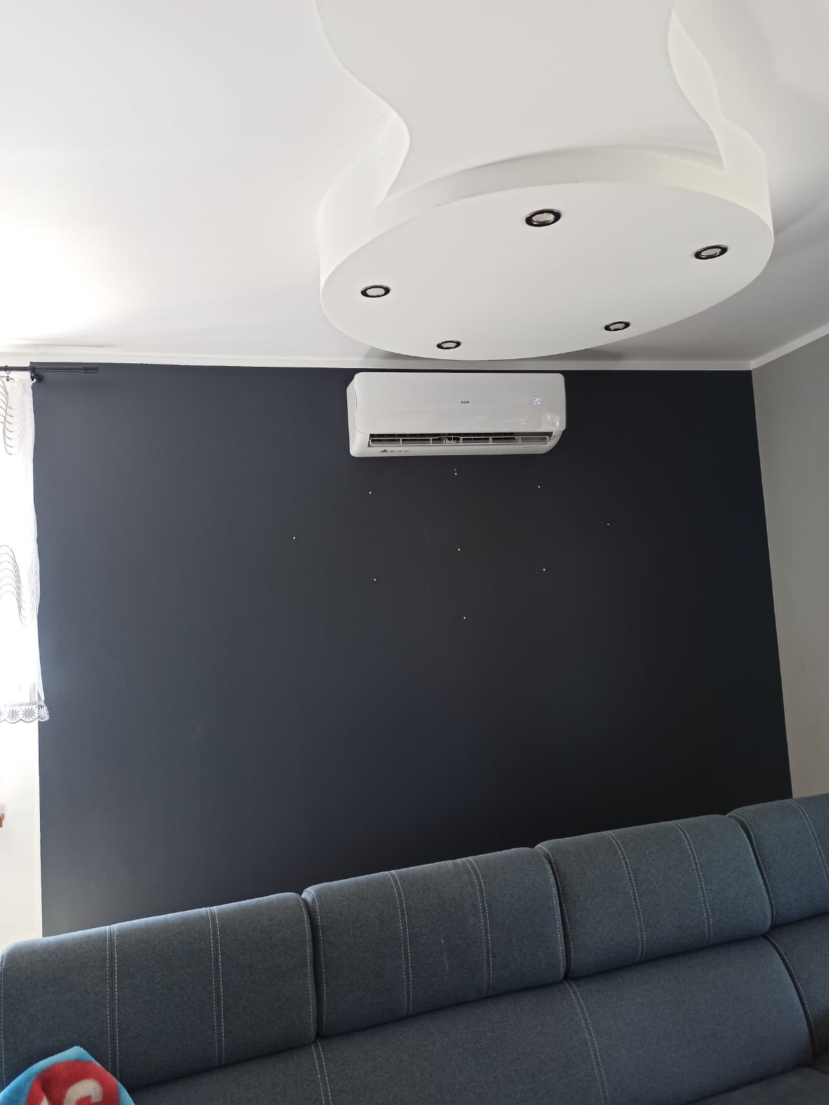 Biały klimatyzator na ciemnoszarej ścianie w salonie, widoczny fragment szarej sofy i okrągła lampa sufitowa z halogenami.