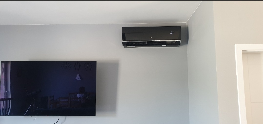 Czarny klimatyzator AUX zamontowany na szarej ścianie nad telewizorem w salonie.