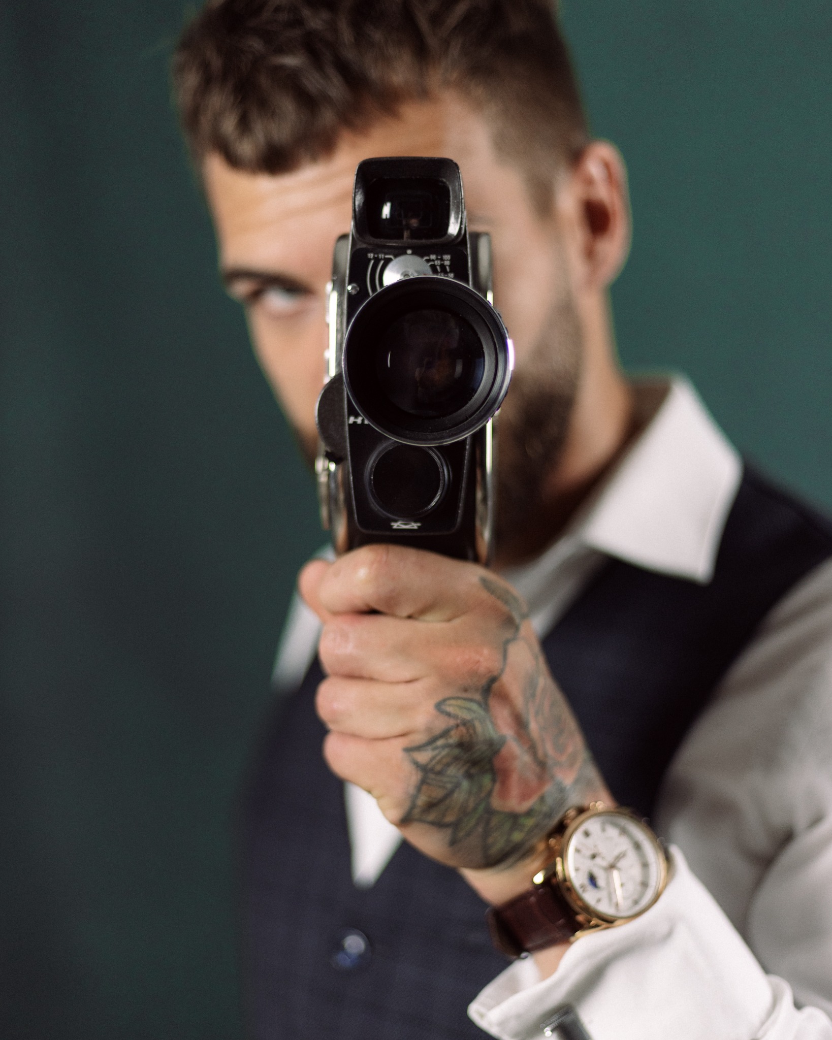 Mężczyzna w kamizelce celuje starą kamerą filmową, widoczny tatuaż na dłoni i zegarek na skórzanym pasku.