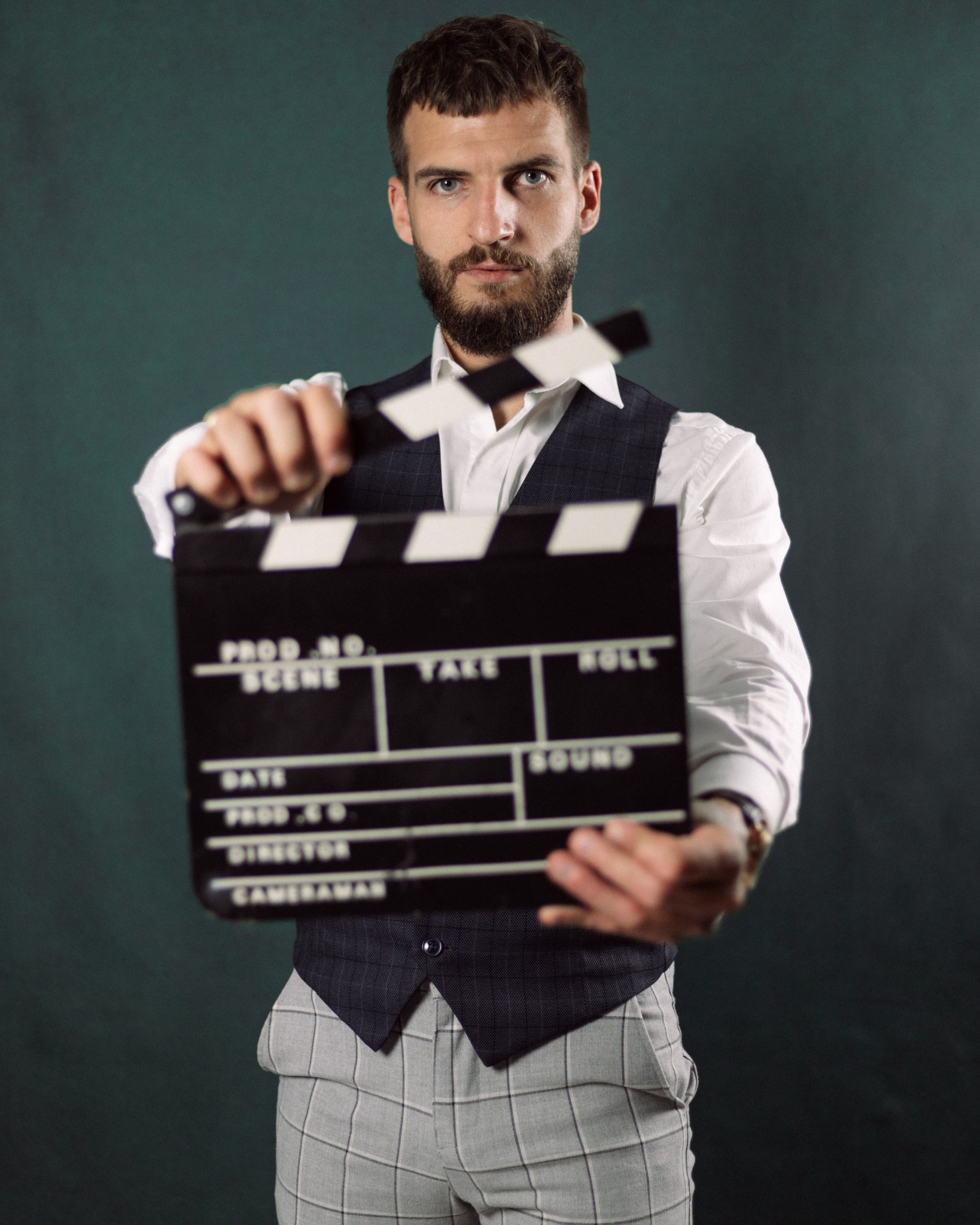 Mężczyzna z brodą, ubrany w białą koszulę i kamizelkę w kratę, trzyma w dłoniach klaps filmowy na tle ciemnozielonym.
