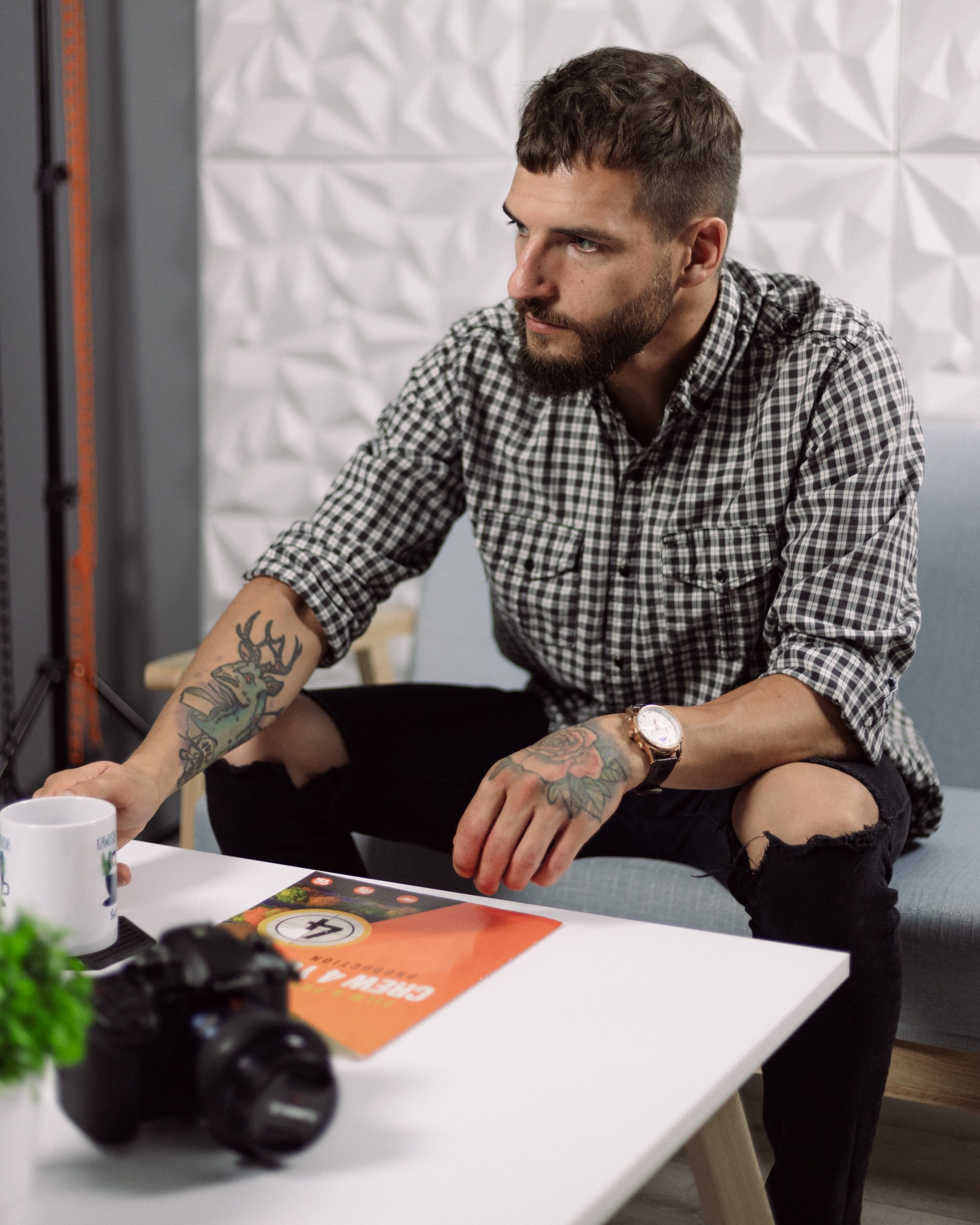 Młody mężczyzna z brodą i tatuażami siedzi na kanapie obok stolika z aparatem, kubkiem i folderem reklamowym 'Crew 4 You Production'.