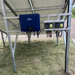 Instalacja fotowoltaiczna: widok od spodu na zamontowane panele, niebieski falownik Hypontech i dwa białe rozłączniki DC SolTEC z ostrzeżeniami o napięciu, na tle trawy.