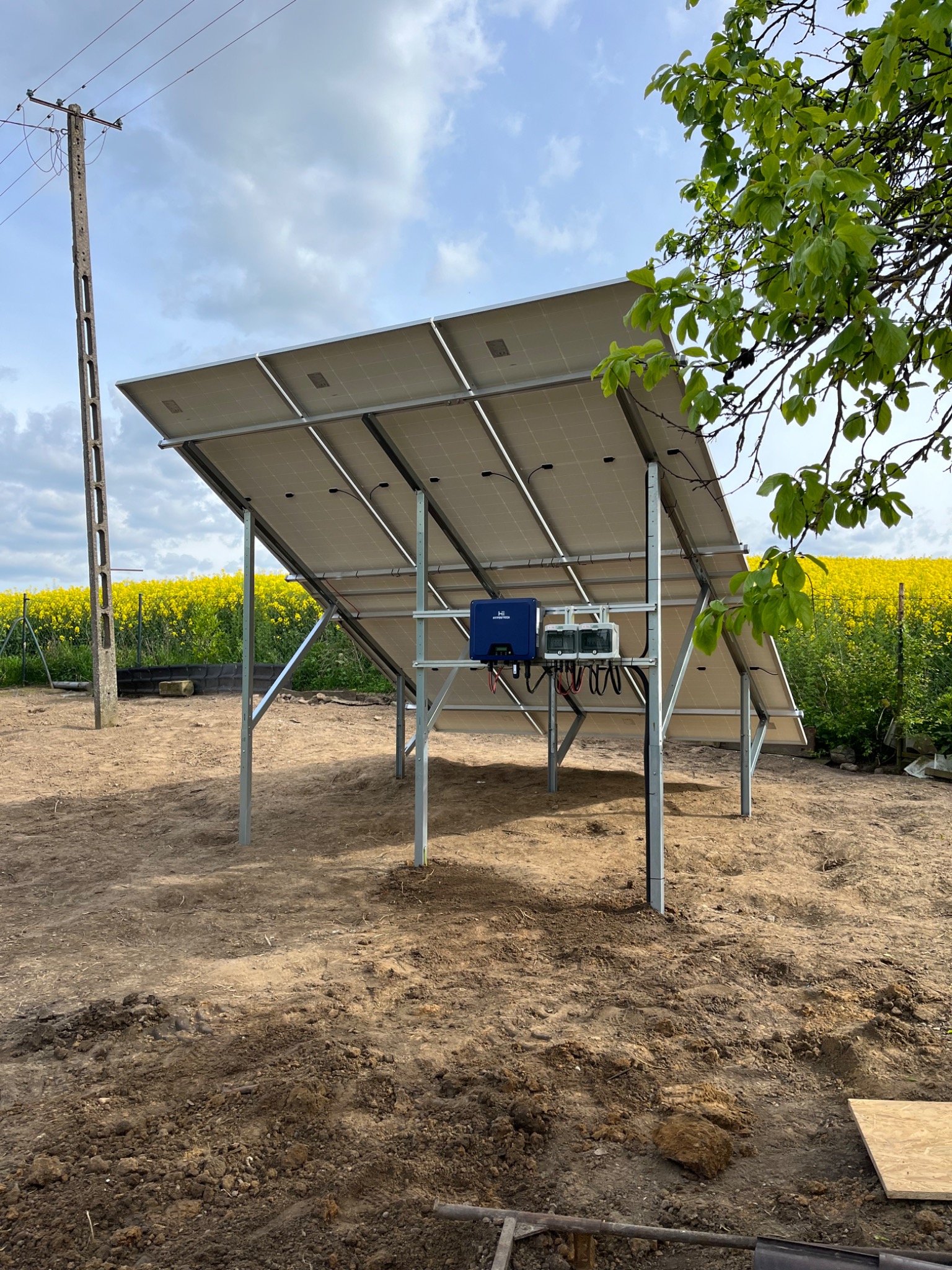 Widok od tyłu na zainstalowane panele słoneczne na metalowej konstrukcji, z inwerterem i przewodami, na tle pola rzepaku i słupa energetycznego.