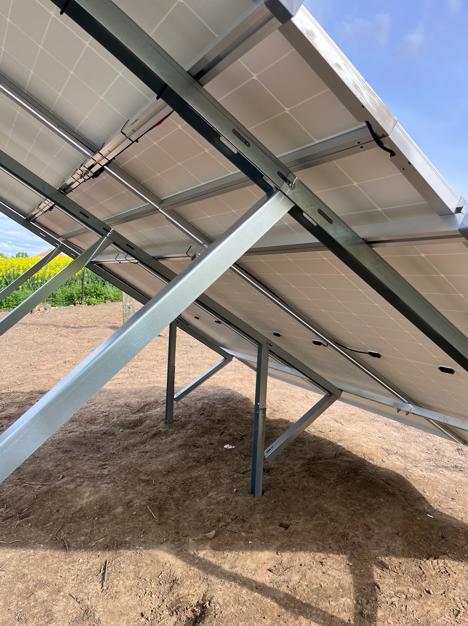 Widok od spodu na panele słoneczne zamontowane na metalowej konstrukcji, widoczne przewody, w tle pole rzepaku i błękitne niebo.