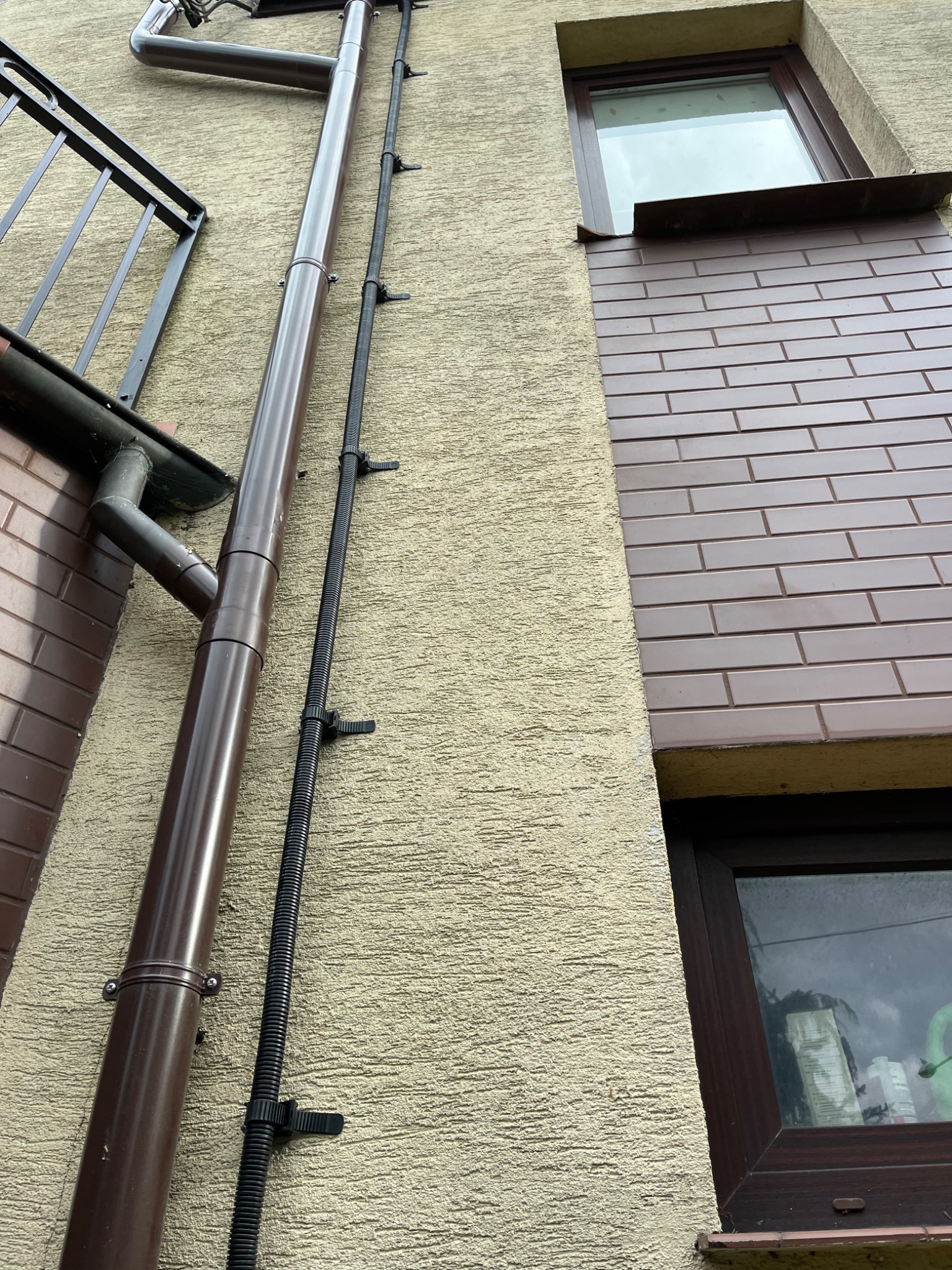 Brązowa rynna spustowa i czarny przewód mocowane do ściany budynku z oknami i balkonem, widok z dołu.