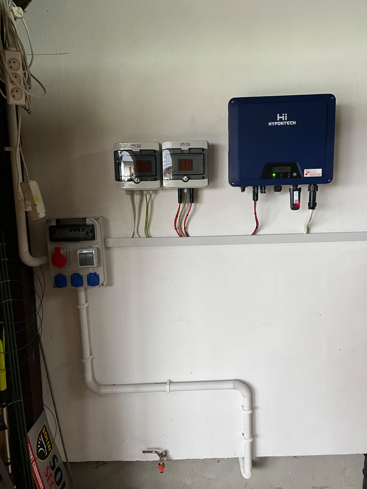 Instalacja fotowoltaiczna: niebieski inwerter Hypdotech zamontowany na białej ścianie obok dwóch rozdzielnic i gniazd siłowych.