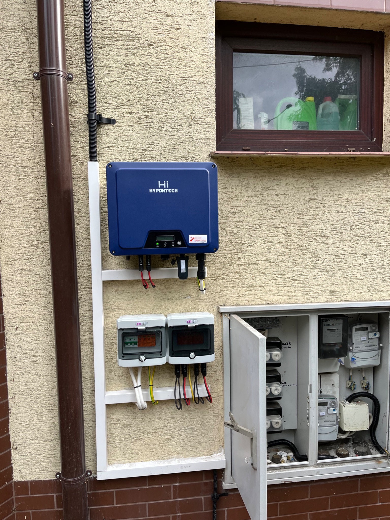 Niebieski falownik HYPONTECH zamontowany na białym stelażu obok brązowej rynny, z widocznymi licznikami i skrzynką elektryczną z otwartymi drzwiczkami na elewacji budynku.