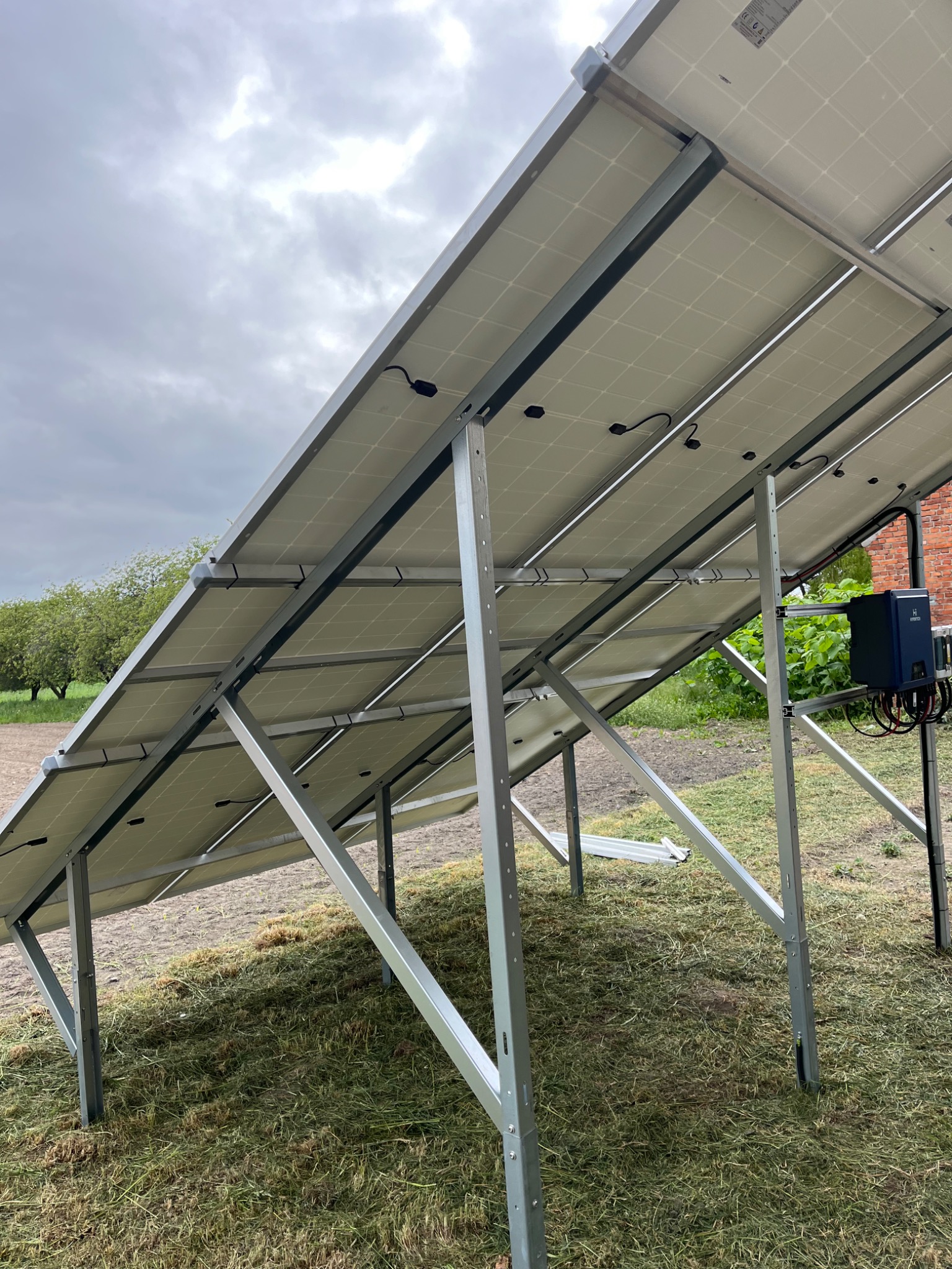 Widok od spodu na panele słoneczne zamontowane na metalowej konstrukcji, widoczne kable i elementy montażowe, tło stanowi trawa i pochmurne niebo.