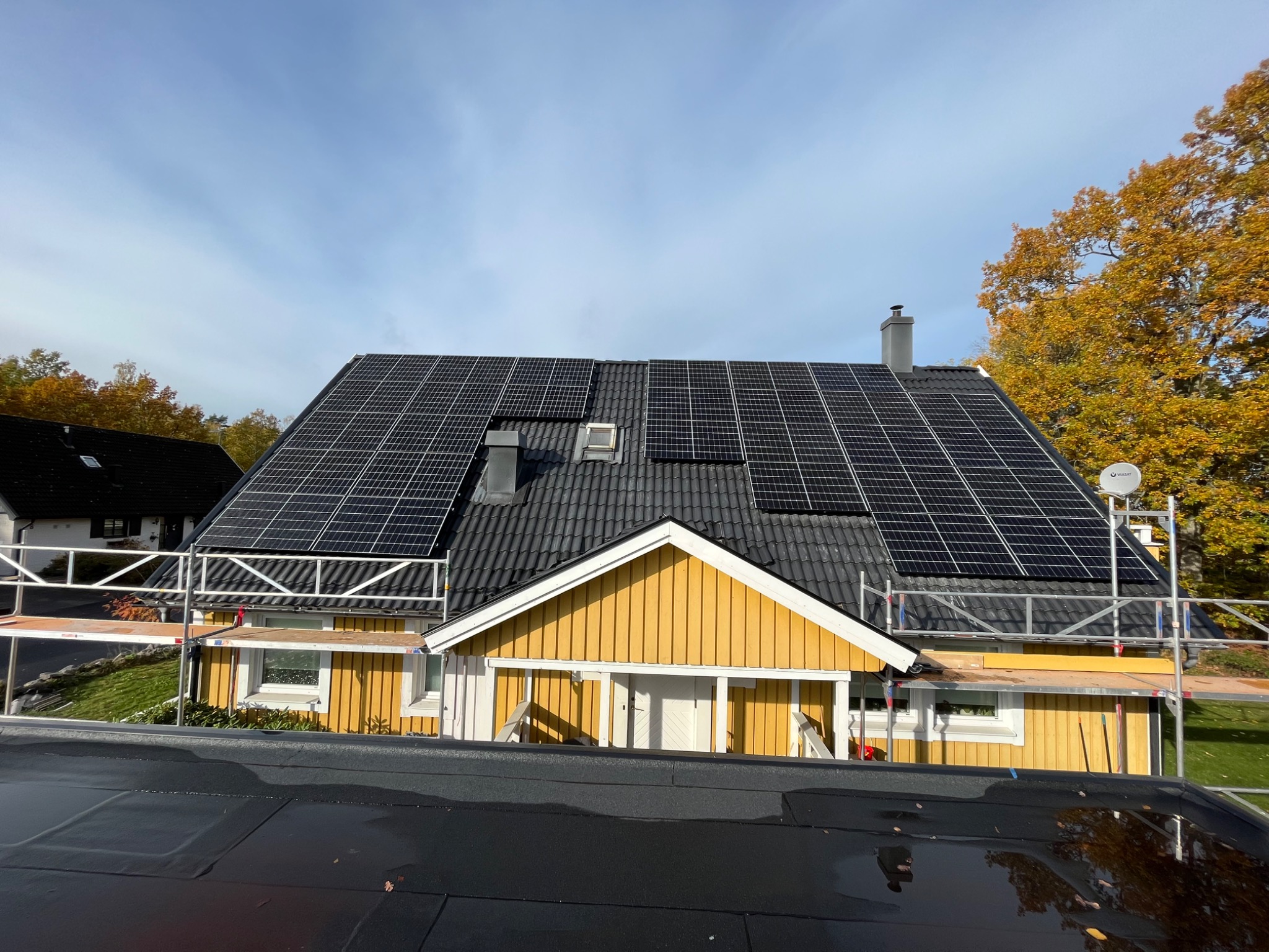 Żółty dom z panelami na dachu, widoczne rusztowania i antena satelitarna, w tle drzewa z jesiennymi liśćmi.