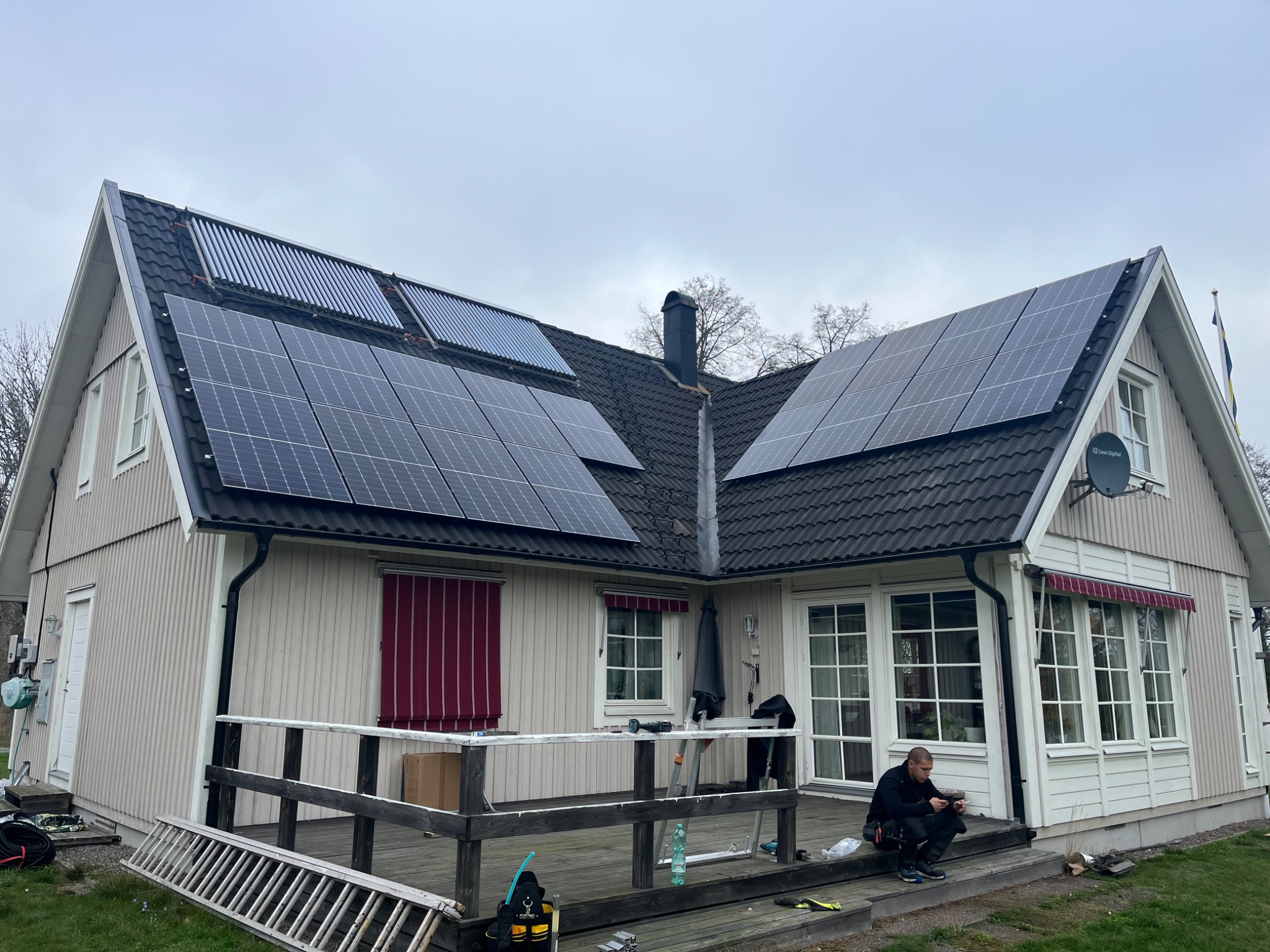 Dom jednorodzinny z panelami słonecznymi i kolektorami słonecznymi na dachu, mężczyzna w roboczym stroju siedzi na tarasie przed domem, obok drabina i torba narzędziowa.