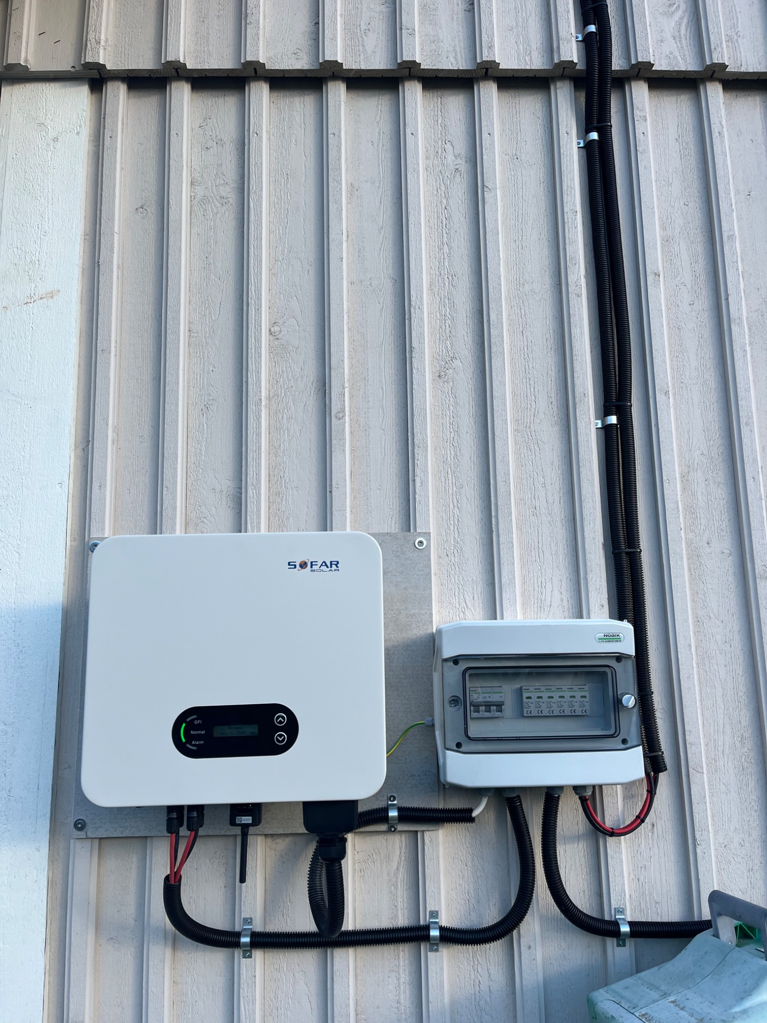 Biały inwerter SOFAR przymocowany do ściany z drewnianych desek obok skrzynki rozdzielczej z bezpiecznikami, widoczne czarne rury karbowane z przewodami.