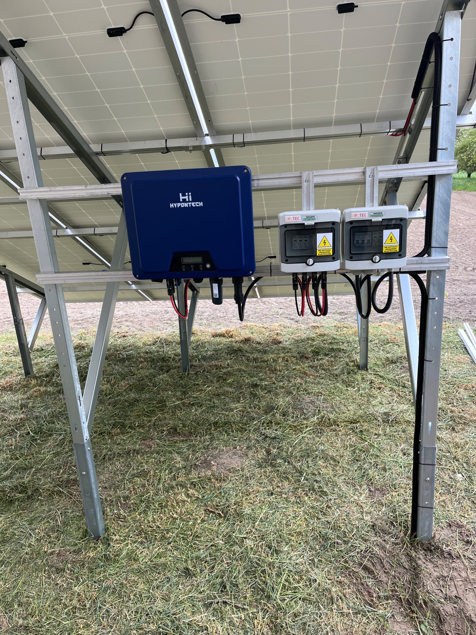 Instalacja fotowoltaiczna: widok od spodu na zamontowane panele, niebieski falownik Hypontech i dwa białe rozłączniki DC SolTEC z ostrzeżeniami o napięciu, na tle trawy.