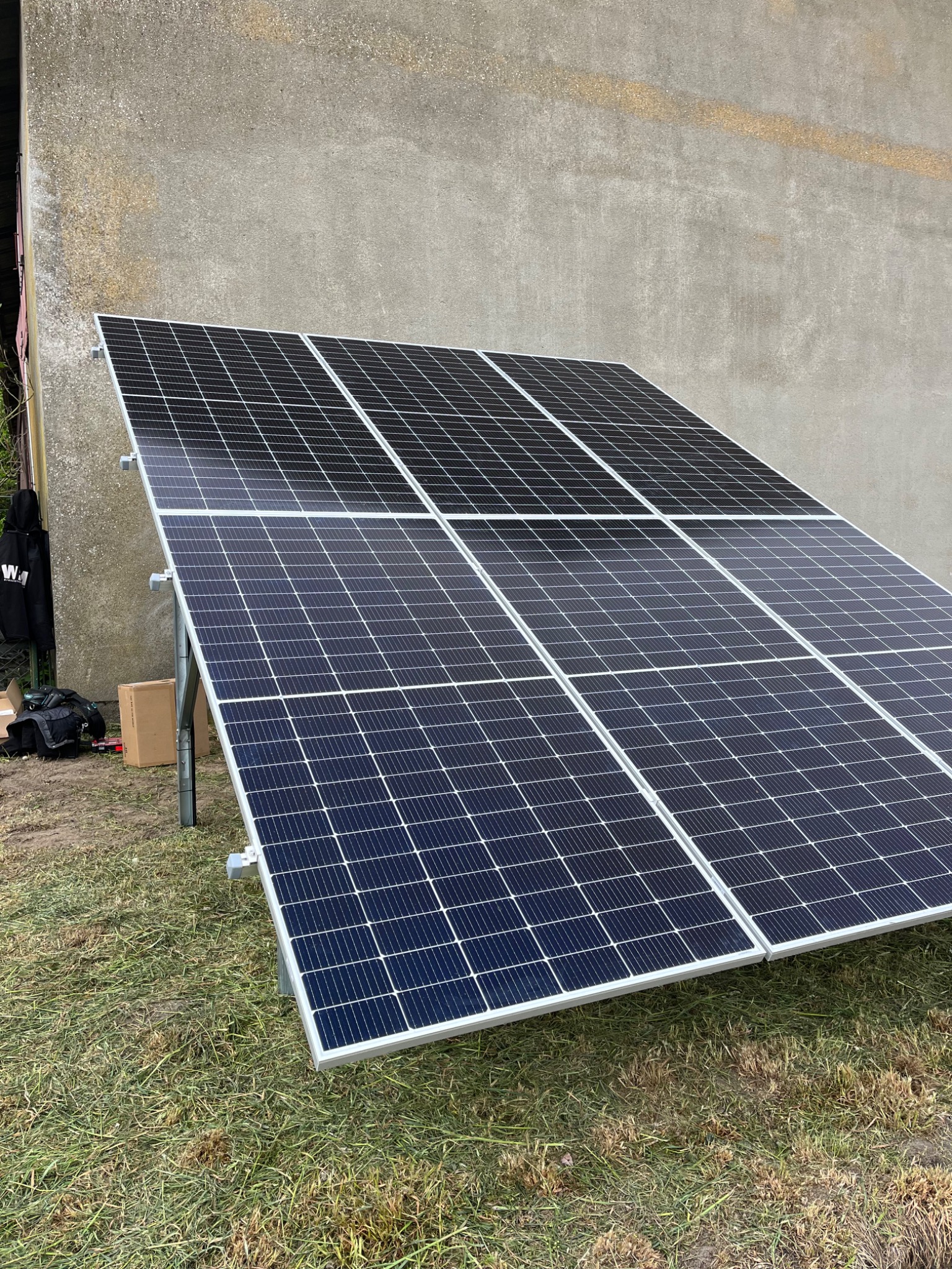 Panel fotowoltaiczny na trawie, na tle szarej ściany budynku, widoczne narzędzia i karton obok paneli.
