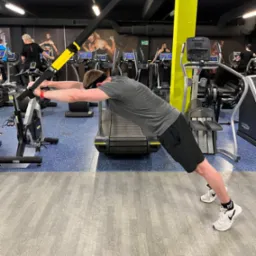 Mężczyzna w szarej koszulce i czarnych spodenkach wykonuje ćwiczenie rozciągające na TRX w siłowni, z widocznym bieżnią, rowerem stacjonarnym i innymi osobami ćwiczącymi w tle.